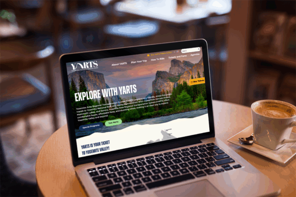 A laptop on a café table displays the YARTS website, featuring a scenic photo of Yosemite Valley; a cup of coffee and a notebook are nearby.