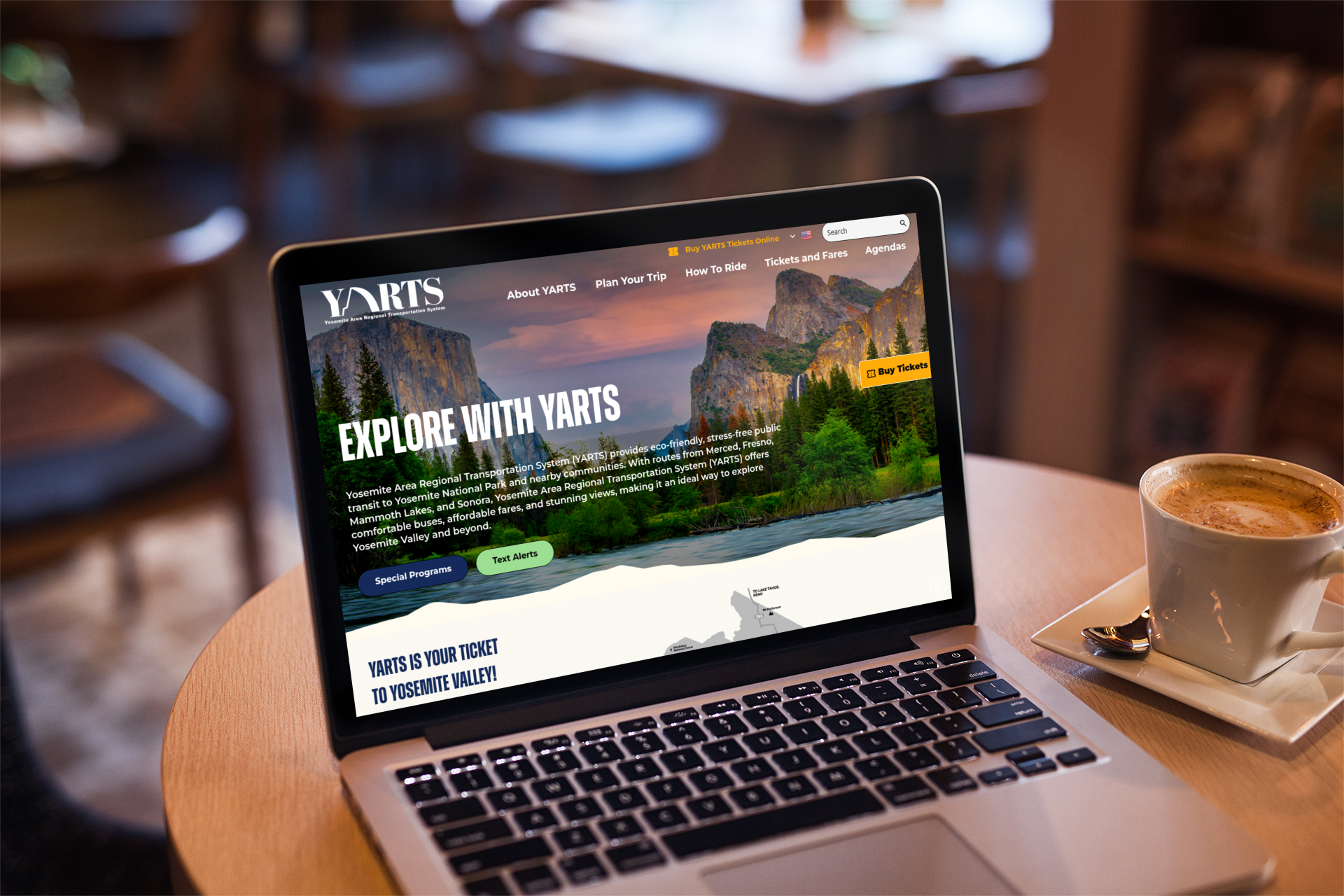 A laptop on a café table displays the YARTS website, featuring a scenic photo of Yosemite Valley; a cup of coffee and a notebook are nearby.