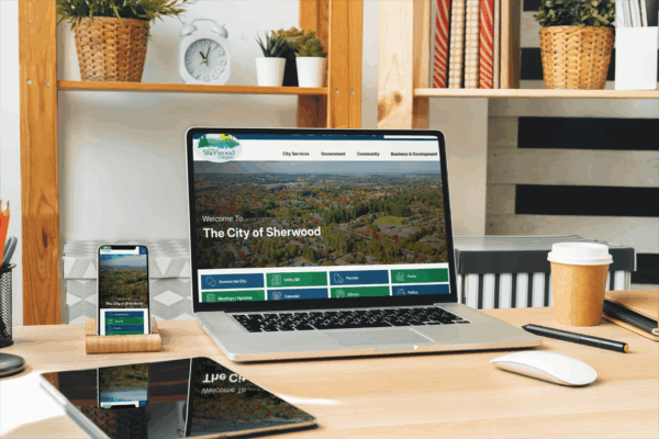 A laptop, smartphone, and tablet on a desk display the City of Sherwood website in a modern office setting with plants and shelves in the background.