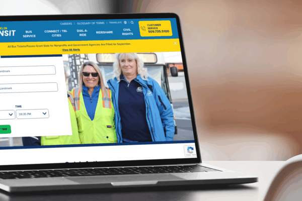 A laptop displays a public transit website showing a route planner form and a photo of two uniformed transit workers standing side by side and smiling.