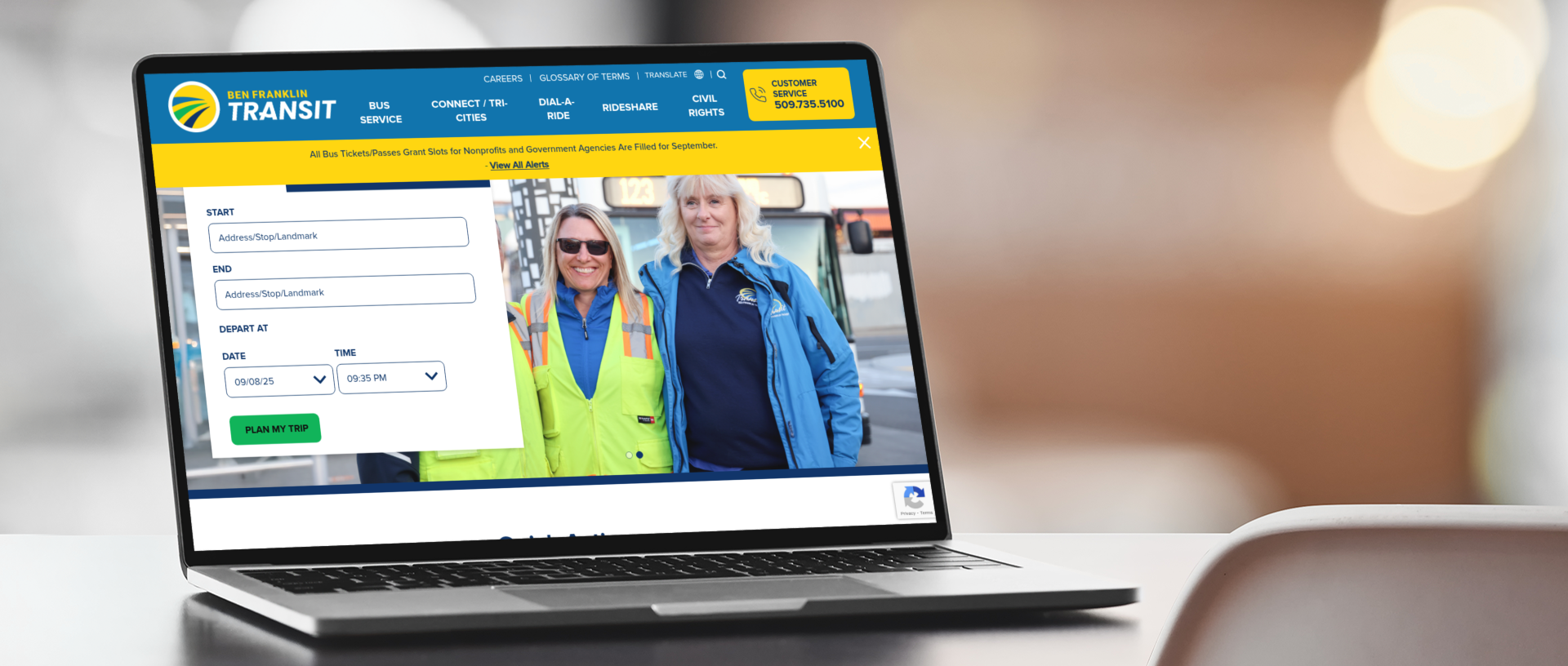 A laptop displays a public transit website showing a route planner form and a photo of two uniformed transit workers standing side by side and smiling.