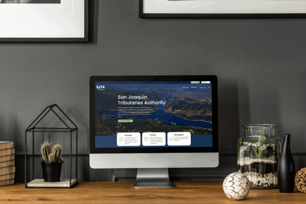 A desktop computer on a wooden desk displays the San Joaquin Tributaries Authority website; books, plants, and decorative items are also on the desk.