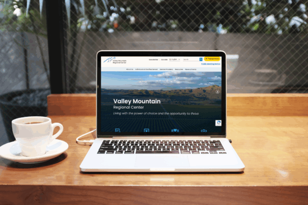 A laptop on a wooden table displays the Valley Mountain Regional Center website; a cup of coffee sits to the left.