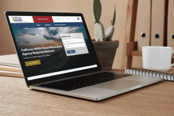 A laptop on a desk displays the California WARN Water/Wastewater Agency Response Network login page. A cactus, spiral notebook, and pen are also on the desk.