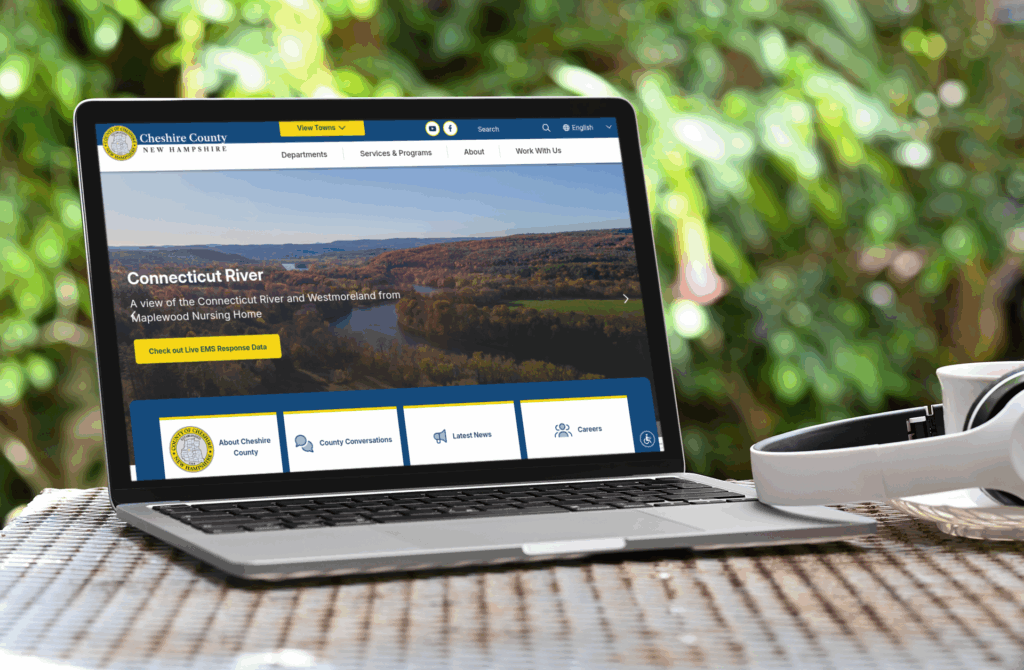 A laptop on a wicker table displays the Cheshire County, New Hampshire website; headphones rest nearby, and trees are visible in the background.