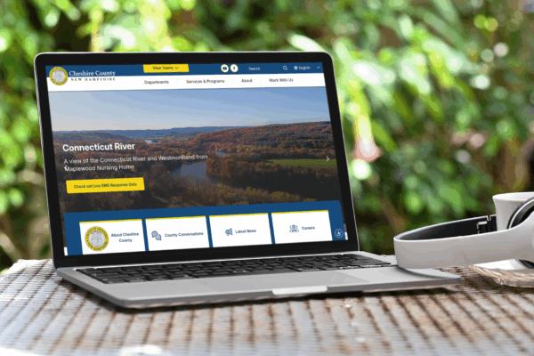 A laptop on a wicker table displays the Cheshire County, New Hampshire website; headphones rest nearby, and trees are visible in the background.