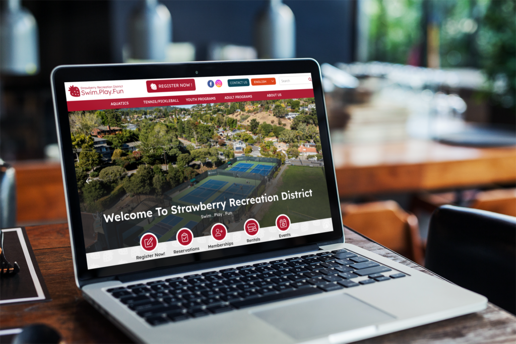 A laptop on a wooden table displays the Strawberry Recreation District website with an aerial view of sports courts and the message, "Welcome To Strawberry Recreation District.