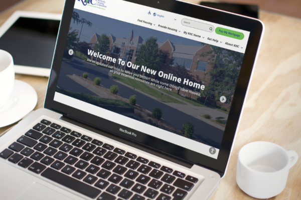 A MacBook Pro displays the homepage of the Kentucky Housing Corporation website, viewed from above next to a cup of coffee and a smartphone on a table.