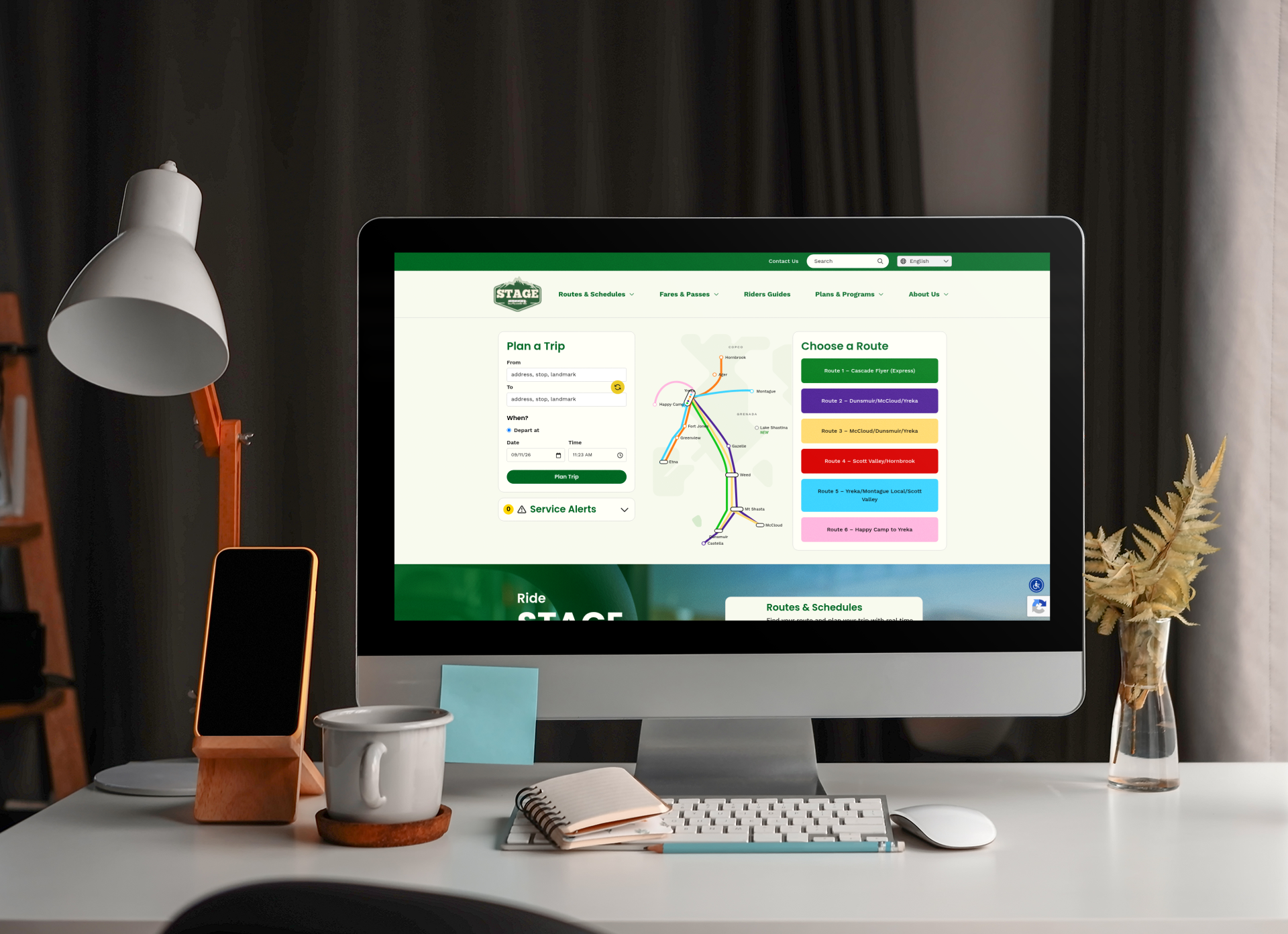 A desktop computer displays a public transit website with route planning options. Office supplies, a cup, and a plant are on the desk in front of the monitor.